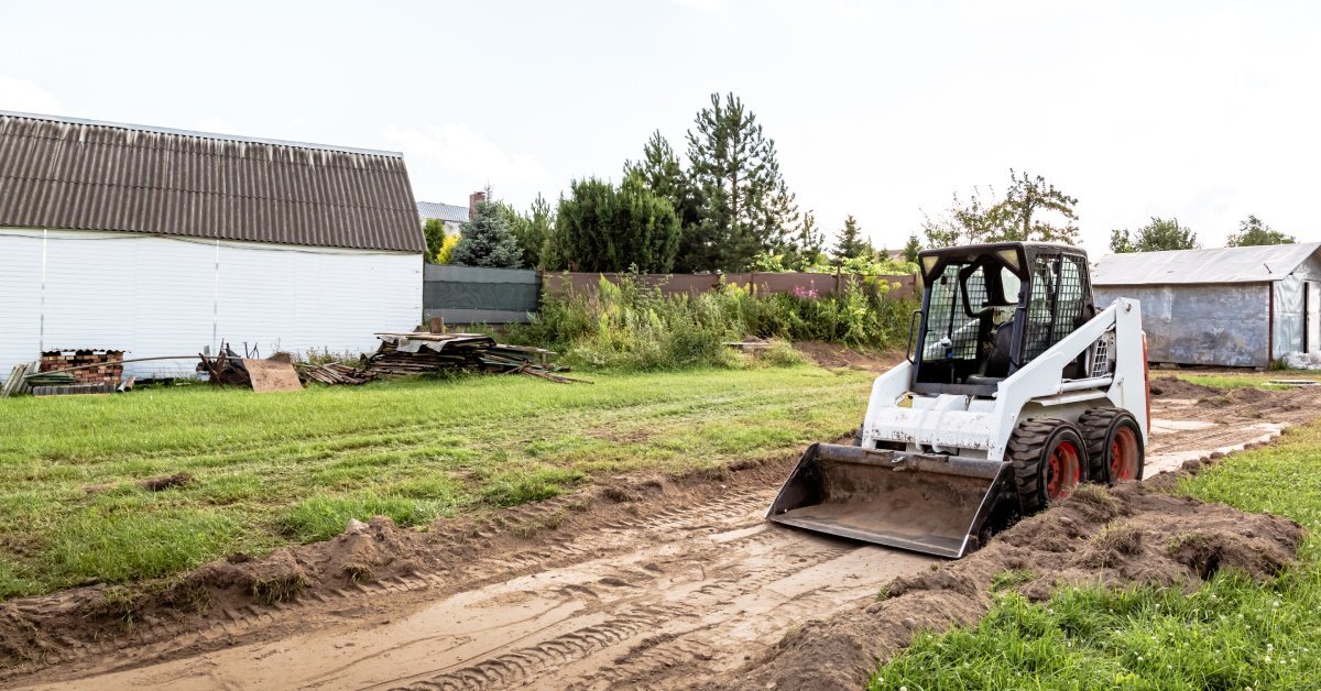 An operator using a skid steer to level ground with a smooth bucket. Grass lies on either side of the path.