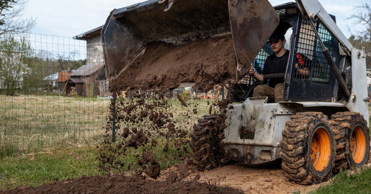 A man operating a skid steer to unload dirt along a flattened path. The smooth bucket attachment has some dirt inside.
